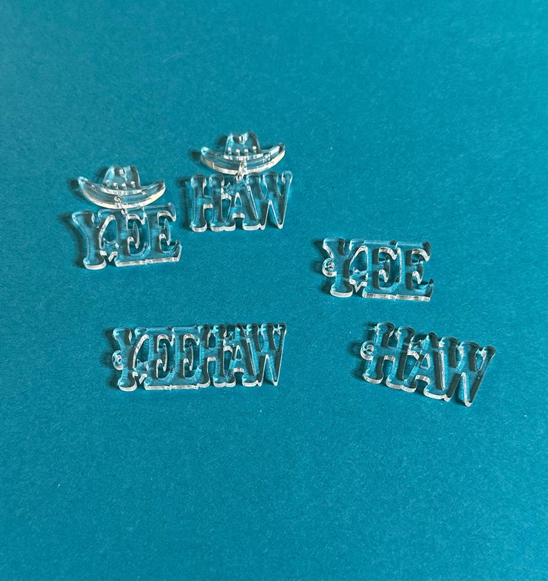 以下が含まれることがあります： 「YEE HAW」の文字とカウボーイハットのデザインが施された透明アクリルカットアウト。カットアウトは様々なサイズと配置で、クラフトや装飾に最適です。背景は無地のティール色です。