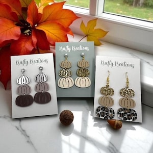 May include: Three pairs of fall-themed earrings on display. Each pair features a pumpkin design, with variations in color and texture, including glitter and leopard print. The cards read "Krafty Kreations".