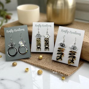 May include: Three pairs of earrings on display. One pair features a diamond ring design, another has the word "BRIDE", and the third says "Mrs". Each pair is on a card with the text "Krafty Kreations".