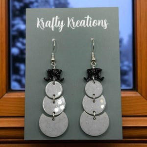 May include: Snowman-themed dangle earrings. Each earring features a black glitter top hat, three light gray circles, and silver-tone hooks. The earrings are displayed on a gray card with the text "Krafty Kreations".