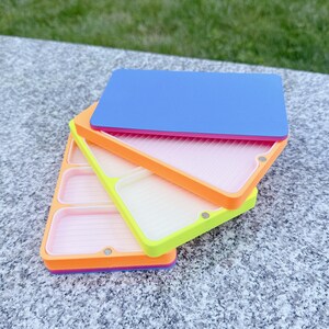 May include: Stack of three colorful craft trays. The top tray is blue, the middle tray is orange, and the bottom tray is yellow. Each tray has a different color border and a pink interior. The trays are stacked on top of each other.