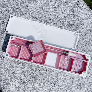 May include: A white and pink organizer with multiple compartments for storing small items. The organizer has a clear lid and a unicorn design on the top. The compartments are filled with small, colorful rhinestones.
