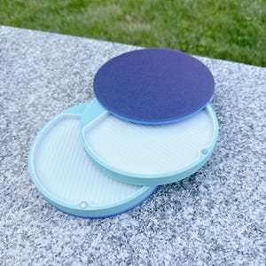 May include: Three-piece, round, stackable tray set. The base trays are light blue with a white interior and a ribbed design. A dark blue, felt-like disc sits on top. The set is designed for organizing small items.