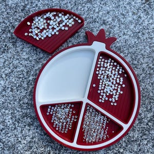 May include: A red and white pomegranate-shaped tray with four compartments for holding small items, such as rhinestones or beads. The tray is filled with clear rhinestones.
