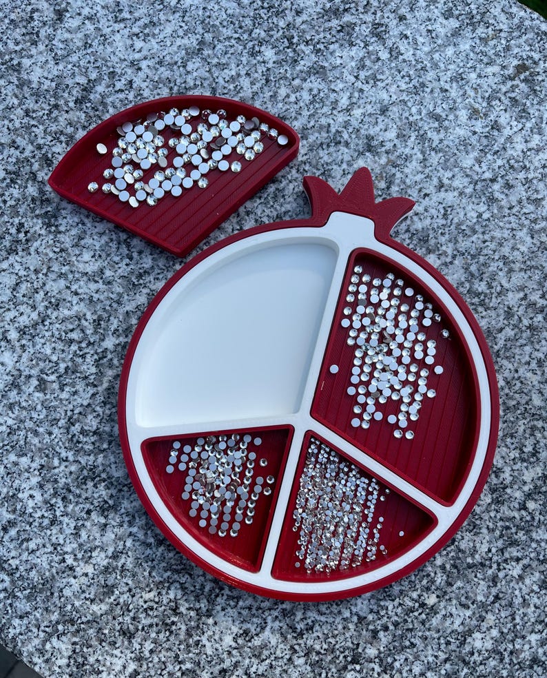 May include: A red and white pomegranate-shaped tray with four compartments for holding small items, such as rhinestones or beads. The tray is filled with clear rhinestones.
