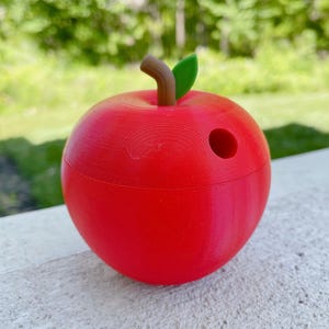 K&ouml;nnte beinhalten: Ein roter, apfelf&ouml;rmiger Bleistifthalter mit einem gr&uuml;nen Blatt und einem braunen Stiel. Der Halter hat oben ein Loch f&uuml;r Bleistifte.