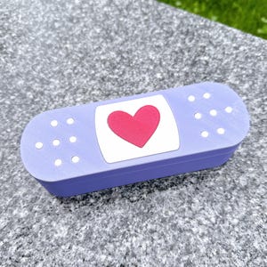 May include: A purple plastic bandaid shaped box with a white heart with a red heart inside.