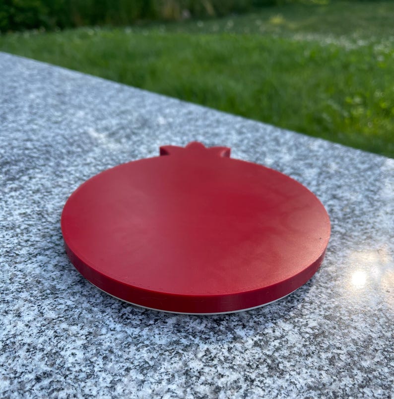 May include: A red, round, pomegranate-shaped coaster with a white rim. The coaster is made of a smooth, hard material and is perfect for protecting surfaces from heat and scratches.