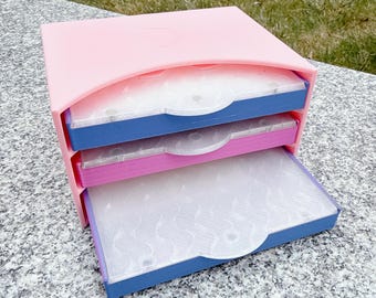 May include: A three-drawer storage organizer in shades of pink, blue, and purple. The top and sides are a soft pink, with three pull-out drawers in blue, pink, and purple. The drawers have a translucent, patterned design.