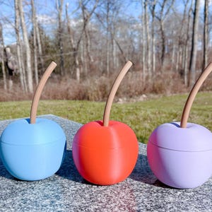 May include: Three ceramic cherry-shaped containers with wooden stems. The containers are blue, red, and purple.
