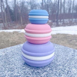 Puede incluir: Una pila de tres recipientes de silicona en forma de macaron de color pastel. El recipiente superior es azul claro, el recipiente del medio es rosa y el recipiente inferior es lavanda.