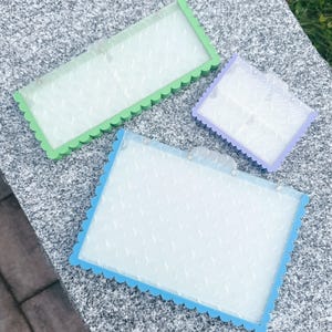 May include: Three decorative picture frames with scalloped edges. The frames are in green, blue, and purple. Each frame has a clear, textured insert. The frames are rectangular and square.