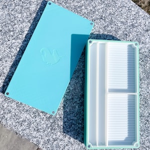 May include: A light blue plastic storage box with a removable lid. The lid has a small, embossed design of a bird. The box has multiple compartments for organizing small items.
