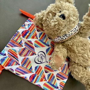Puede incluir: Un oso de peluche con un collar decorativo plateado y dorado descansa junto a una bolsa con cord&oacute;n. La bolsa presenta un fondo blanco con patrones de corazones a rayas de colores y un monograma azul marino.
