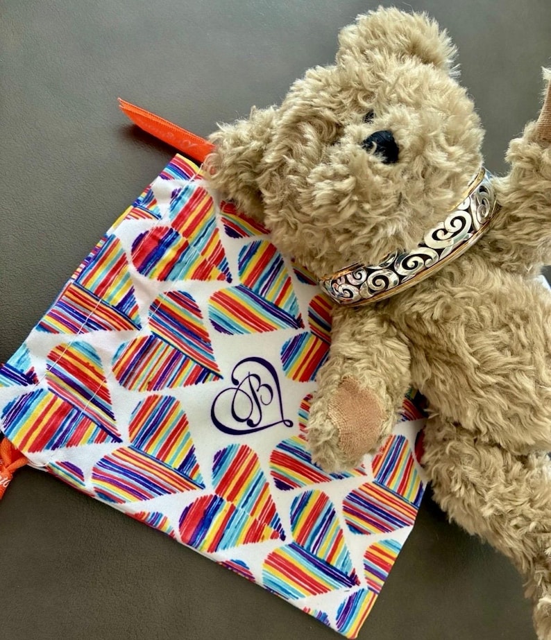 Puede incluir: Un oso de peluche con un collar decorativo plateado y dorado descansa junto a una bolsa con cord&oacute;n. La bolsa presenta un fondo blanco con patrones de corazones a rayas de colores y un monograma azul marino.