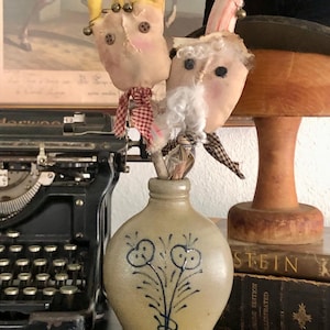 May include: A tan stoneware vase with a blue floral design holds two handmade fabric dolls. The dolls have button eyes, rosy cheeks, and fabric crowns or hats. A vintage typewriter and books are in the background.