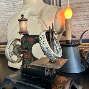 Puede incluir: Una m&aacute;quina de coser negra vintage con detalles florales y una pulsera de plata y oro. La m&aacute;quina tiene un carrete de hilo de madera y una rueda decorativa. Una vela encendida y un maniqu&iacute; est&aacute;n en el fondo.