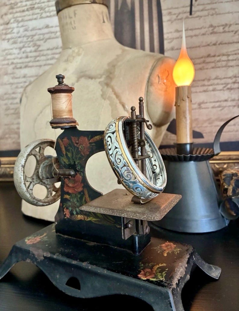 Puede incluir: Una m&aacute;quina de coser negra vintage con detalles florales y una pulsera de plata y oro. La m&aacute;quina tiene un carrete de hilo de madera y una rueda decorativa. Una vela encendida y un maniqu&iacute; est&aacute;n en el fondo.