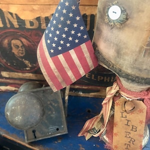 May include: A patriotic still life featuring a small American flag with red and white stripes and white stars. A vintage doll with a button eye and a tag that says "LIBERTY" is also present. An antique door knob and lock are visible.