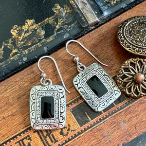 May include: Silver-tone rectangular earrings with a black center stone and a border of small, clear crystals. The earrings have a decorative, engraved frame and a hook closure. Two decorative buttons are also visible.