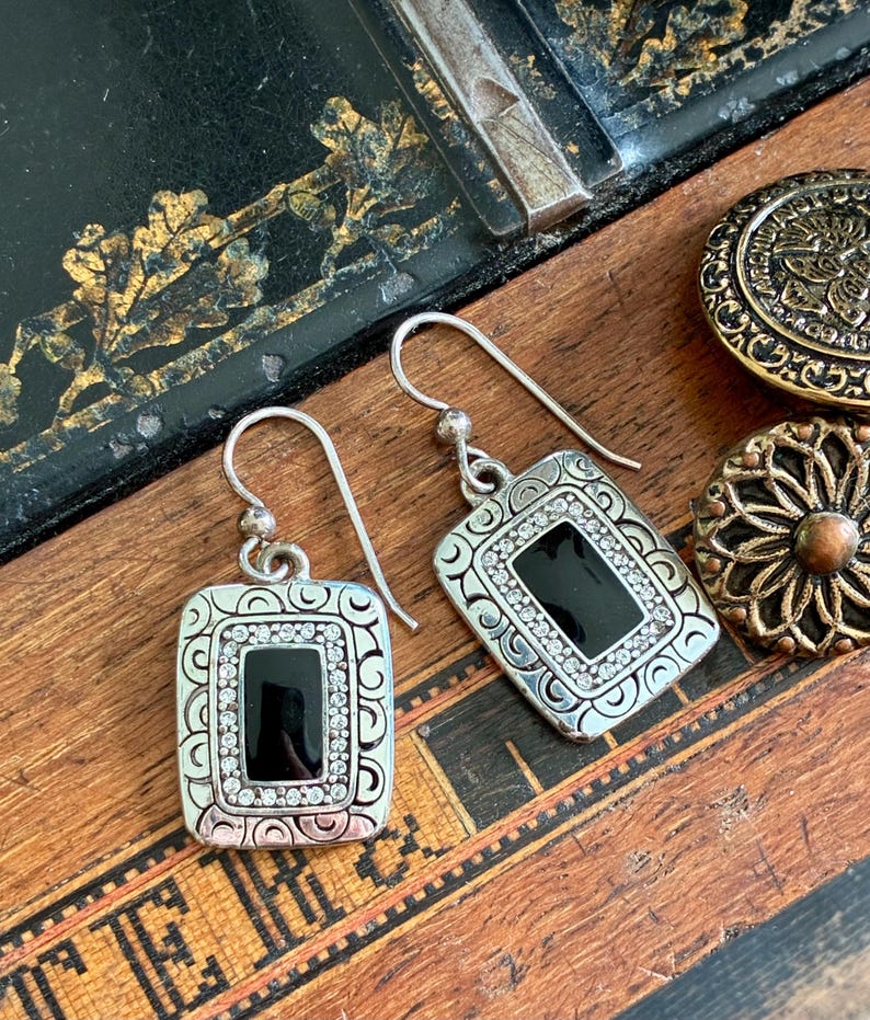 May include: Silver-tone rectangular earrings with a black center stone and a border of small, clear crystals. The earrings have a decorative, engraved frame and a hook closure. Two decorative buttons are also visible.