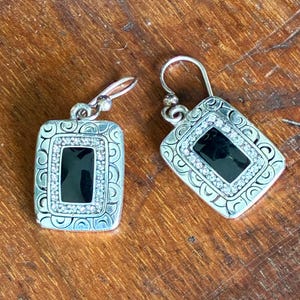 May include: Silver-toned dangle earrings with a rectangular black center stone, surrounded by a row of small, clear crystals. The earrings have a decorative, patterned border and a simple hook closure.