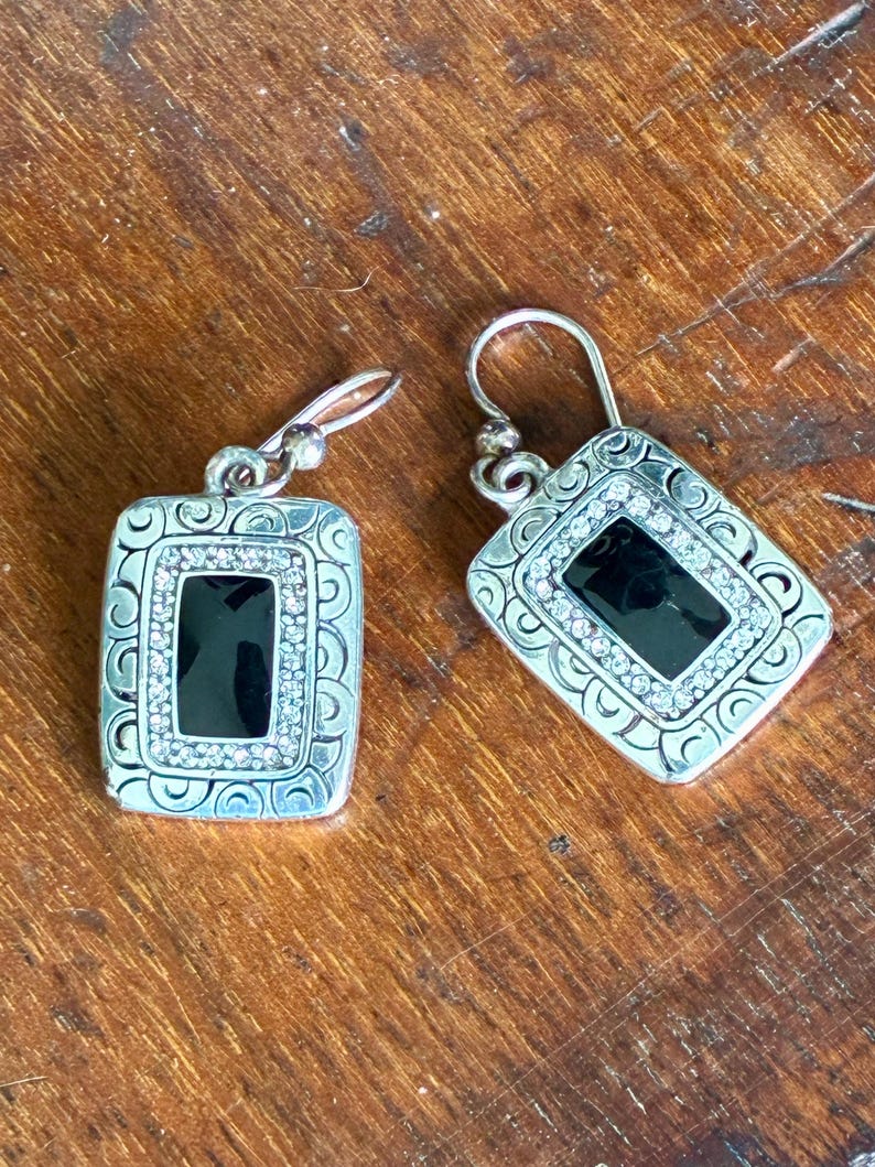 May include: Silver-toned dangle earrings with a rectangular black center stone, surrounded by a row of small, clear crystals. The earrings have a decorative, patterned border and a simple hook closure.