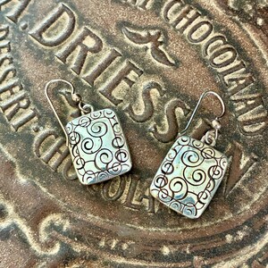 May include: A pair of silver-toned rectangular earrings with a swirling design. The earrings have a hook closure and are set against a brown, textured surface with the words "DESSERT CHOCOLATE ADRIES" embossed on it.