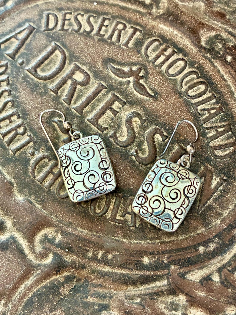 May include: A pair of silver-toned rectangular earrings with a swirling design. The earrings have a hook closure and are set against a brown, textured surface with the words "DESSERT CHOCOLATE ADRIES" embossed on it.