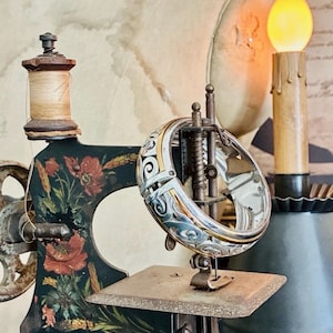 Puede incluir: Una m&aacute;quina de coser vintage con base negra y detalles florales, con una pulsera plateada y dorada. La m&aacute;quina tiene un carrete de hilo de madera y una vela decorativa en el fondo. La pulsera tiene un di&aacute;metro de aproximadamente 5 cm.