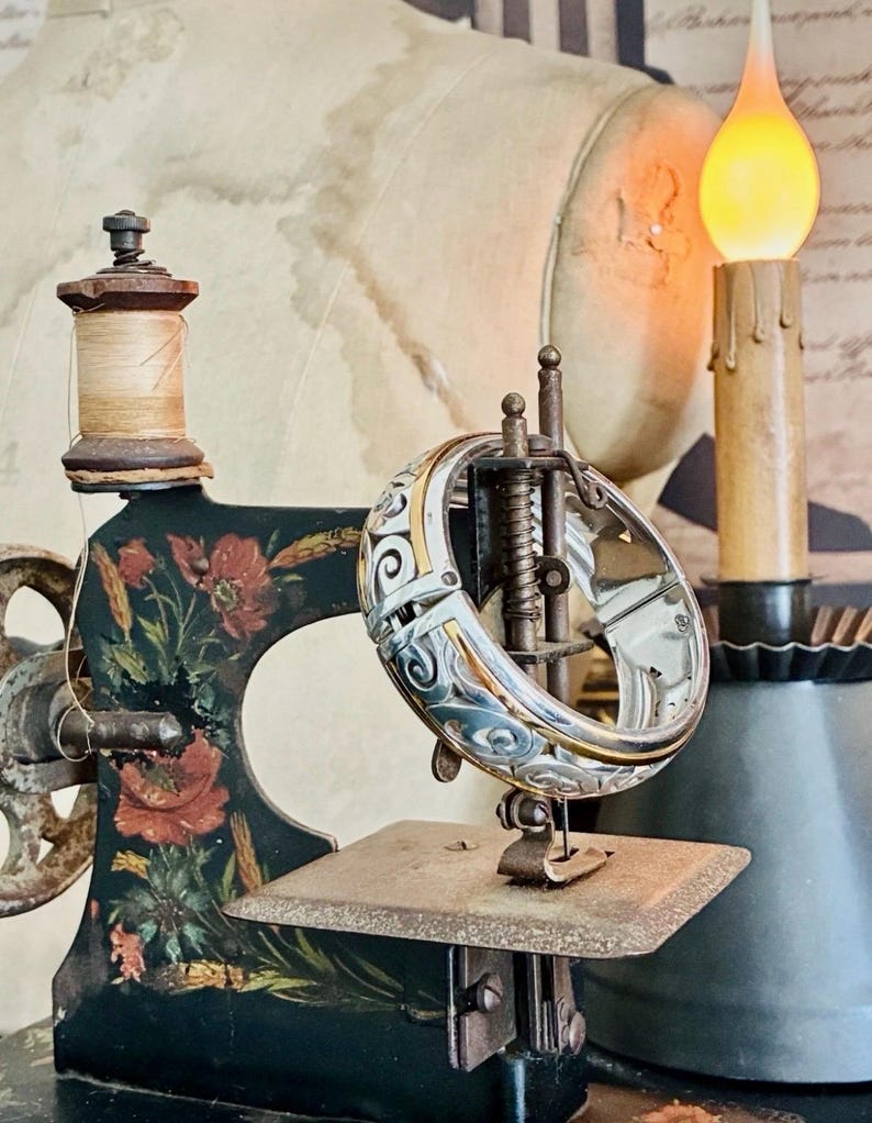 Puede incluir: Una m&aacute;quina de coser vintage con base negra y detalles florales, con una pulsera plateada y dorada. La m&aacute;quina tiene un carrete de hilo de madera y una vela decorativa en el fondo. La pulsera tiene un di&aacute;metro de aproximadamente 5 cm.