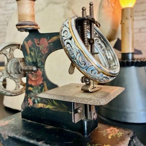 Puede incluir: Una antigua m&aacute;quina de coser negra con detalles florales y una pulsera decorativa de plata y oro. La pulsera se exhibe en el &aacute;rea de la aguja de la m&aacute;quina. Un carrete de hilo se encuentra en la parte superior.