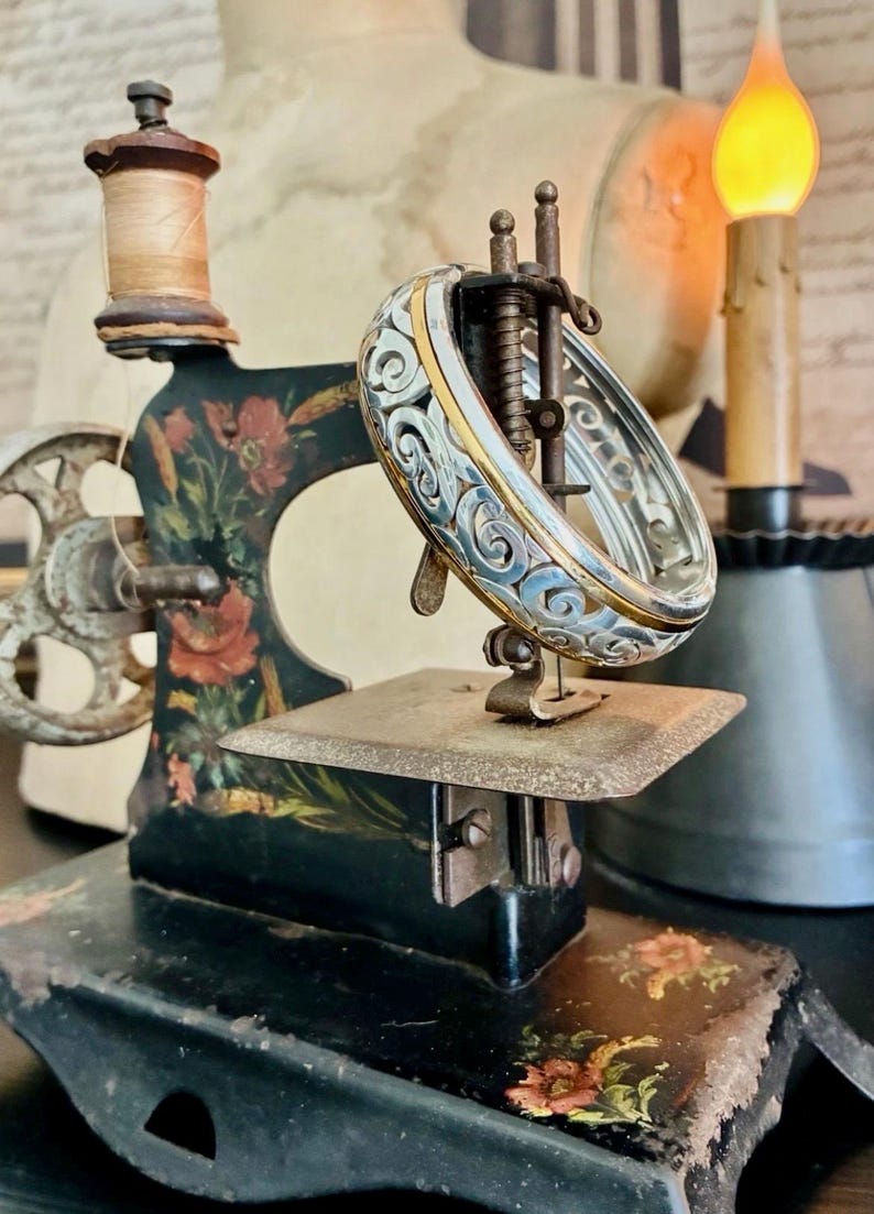 Puede incluir: Una antigua m&aacute;quina de coser negra con detalles florales y una pulsera decorativa de plata y oro. La pulsera se exhibe en el &aacute;rea de la aguja de la m&aacute;quina. Un carrete de hilo se encuentra en la parte superior.