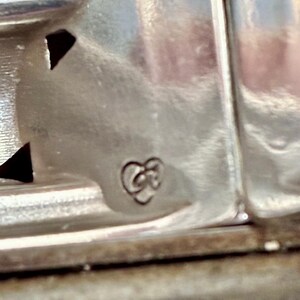Puede incluir: Primer plano de un objeto de metal plateado con un dise&ntilde;o decorativo. El objeto tiene una superficie texturizada y un peque&ntilde;o emblema estilizado en forma de coraz&oacute;n. La imagen est&aacute; bien iluminada, destacando los detalles del trabajo en metal.