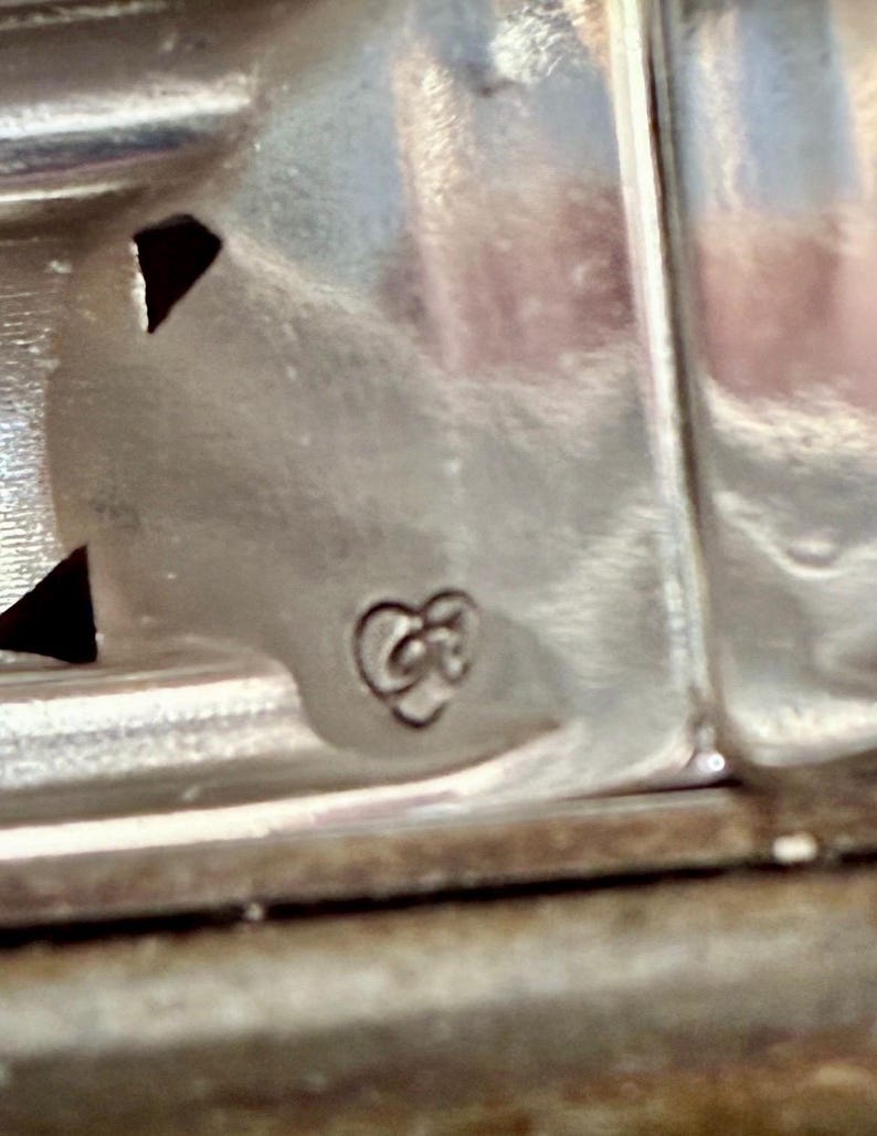 Puede incluir: Primer plano de un objeto de metal plateado con un dise&ntilde;o decorativo. El objeto tiene una superficie texturizada y un peque&ntilde;o emblema estilizado en forma de coraz&oacute;n. La imagen est&aacute; bien iluminada, destacando los detalles del trabajo en metal.