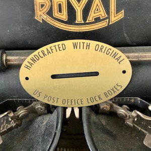 May include: A gold-colored metal plate with the text "HANDCRAFTED WITH ORIGINAL US POST OFFICE LOCK BOXES" and a slot. The plate is set against a black background with the word "ROYAL" in gold lettering.