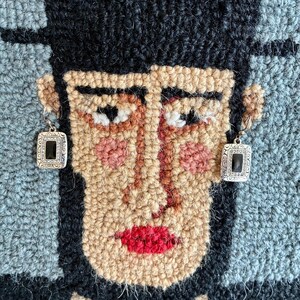 May include: A close-up of a rug with a face design. The face is made of various yarn colors, including tan, black, red, and pink. The person is wearing silver earrings with black square stones. The background is blue and black.