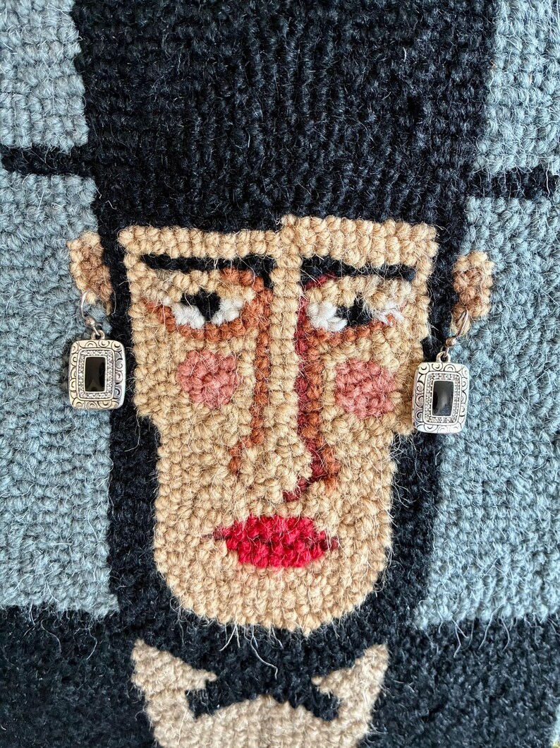May include: A close-up of a rug with a face design. The face is made of various yarn colors, including tan, black, red, and pink. The person is wearing silver earrings with black square stones. The background is blue and black.