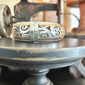 Puede incluir: Pulsera articulada plateada con detalles dorados. La pulsera presenta un dise&ntilde;o calado intrincado con patrones arremolinados. Los detalles dorados son visibles en los bordes superior e inferior. La pulsera se exhibe sobre una superficie oscura y redonda.