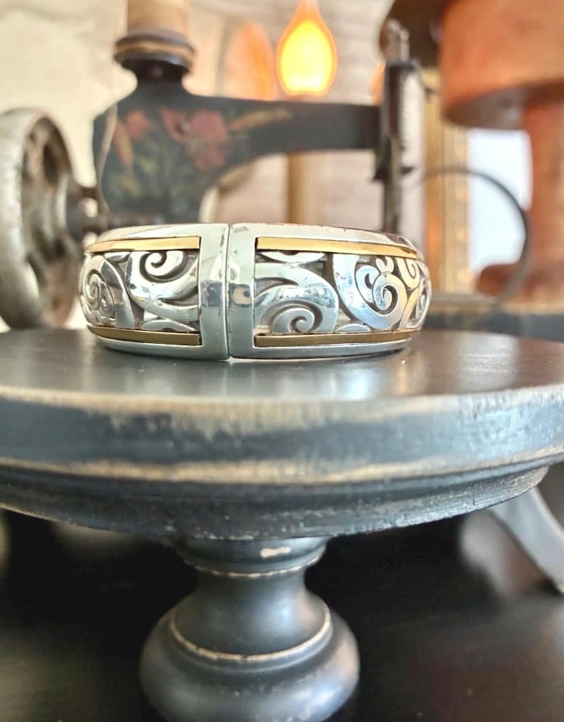 Puede incluir: Pulsera articulada plateada con detalles dorados. La pulsera presenta un dise&ntilde;o calado intrincado con patrones arremolinados. Los detalles dorados son visibles en los bordes superior e inferior. La pulsera se exhibe sobre una superficie oscura y redonda.