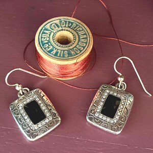 May include: A pair of silver-toned earrings with a rectangular black center stone, surrounded by small clear crystals and decorative scrollwork. The earrings are displayed with a vintage wooden spool of red thread.