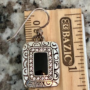 May include: Silver-toned rectangular earring with a black center and a border of small, clear crystals. The earring has a decorative, swirling pattern around the edges and a hook for pierced ears. The earring is approximately 1 inch long.