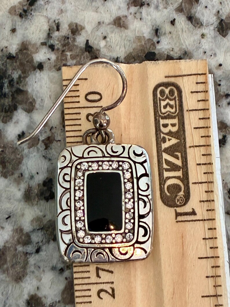 May include: Silver-toned rectangular earring with a black center and a border of small, clear crystals. The earring has a decorative, swirling pattern around the edges and a hook for pierced ears. The earring is approximately 1 inch long.