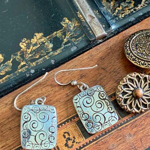 May include: Silver rectangular earrings with a swirling design. The earrings have a hook closure and are displayed on a wooden surface with antique buttons and a sewing machine part.