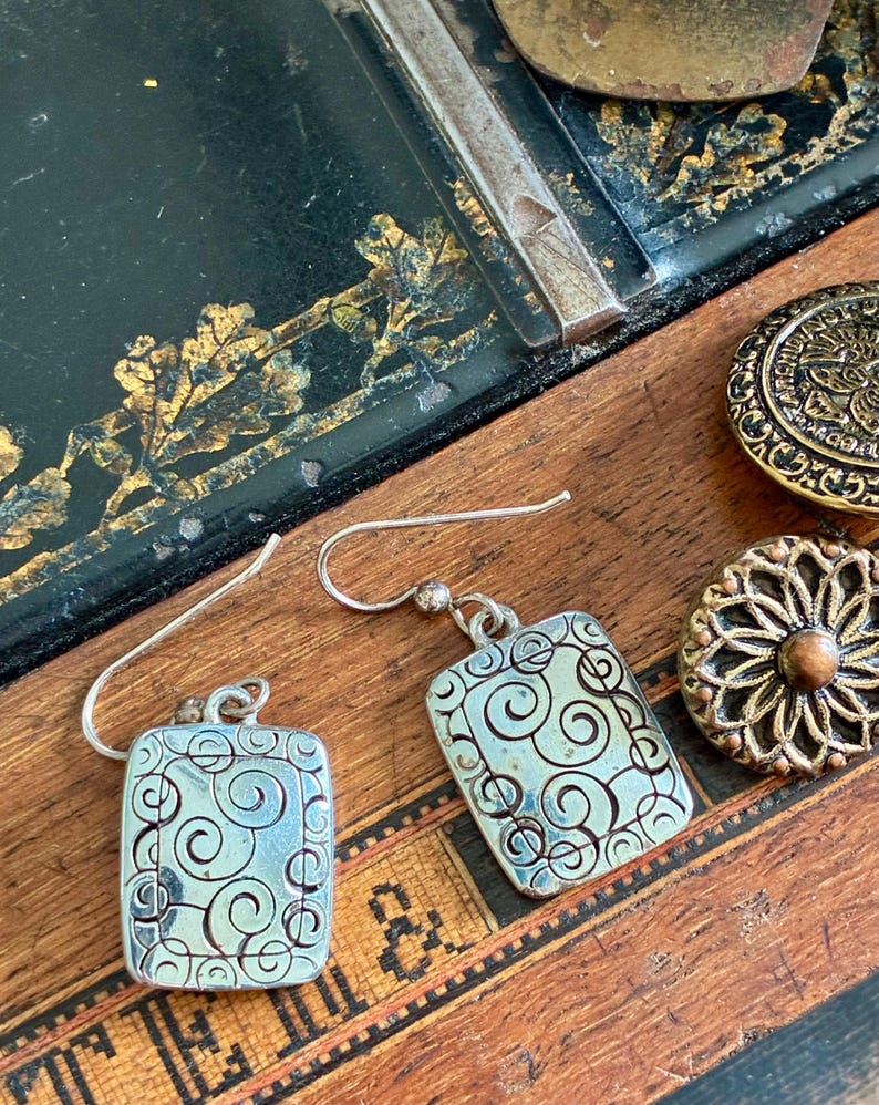 May include: Silver rectangular earrings with a swirling design. The earrings have a hook closure and are displayed on a wooden surface with antique buttons and a sewing machine part.