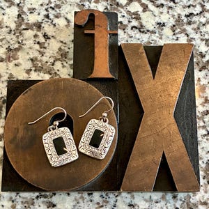 May include: A pair of silver-toned earrings with a rectangular black center and decorative border. The earrings are displayed on a dark brown circular object, with a copper-colored letter "f" and a wooden letter "X" in the background.