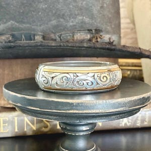 Puede incluir: Un brazalete plateado y dorado con un intrincado dise&ntilde;o de remolinos. El brazalete se exhibe sobre un peque&ntilde;o pedestal de madera negro y desgastado. Un sombrero vintage se encuentra en el fondo.
