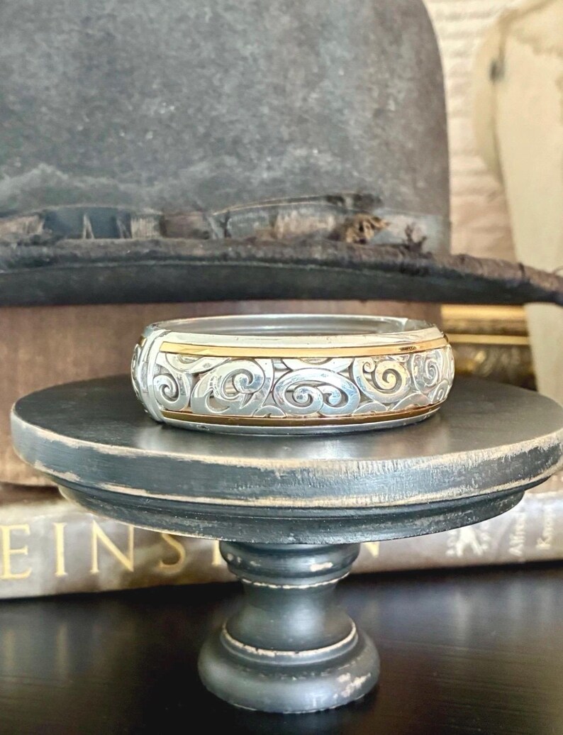 Puede incluir: Un brazalete plateado y dorado con un intrincado dise&ntilde;o de remolinos. El brazalete se exhibe sobre un peque&ntilde;o pedestal de madera negro y desgastado. Un sombrero vintage se encuentra en el fondo.