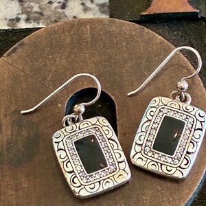May include: A pair of silver-toned earrings with a rectangular black center and a border of small, clear crystals. The earrings have a decorative, patterned frame and a hook closure.