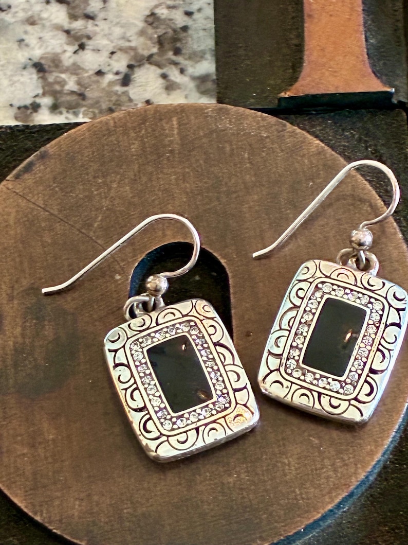 May include: A pair of silver-toned earrings with a rectangular black center and a border of small, clear crystals. The earrings have a decorative, patterned frame and a hook closure.